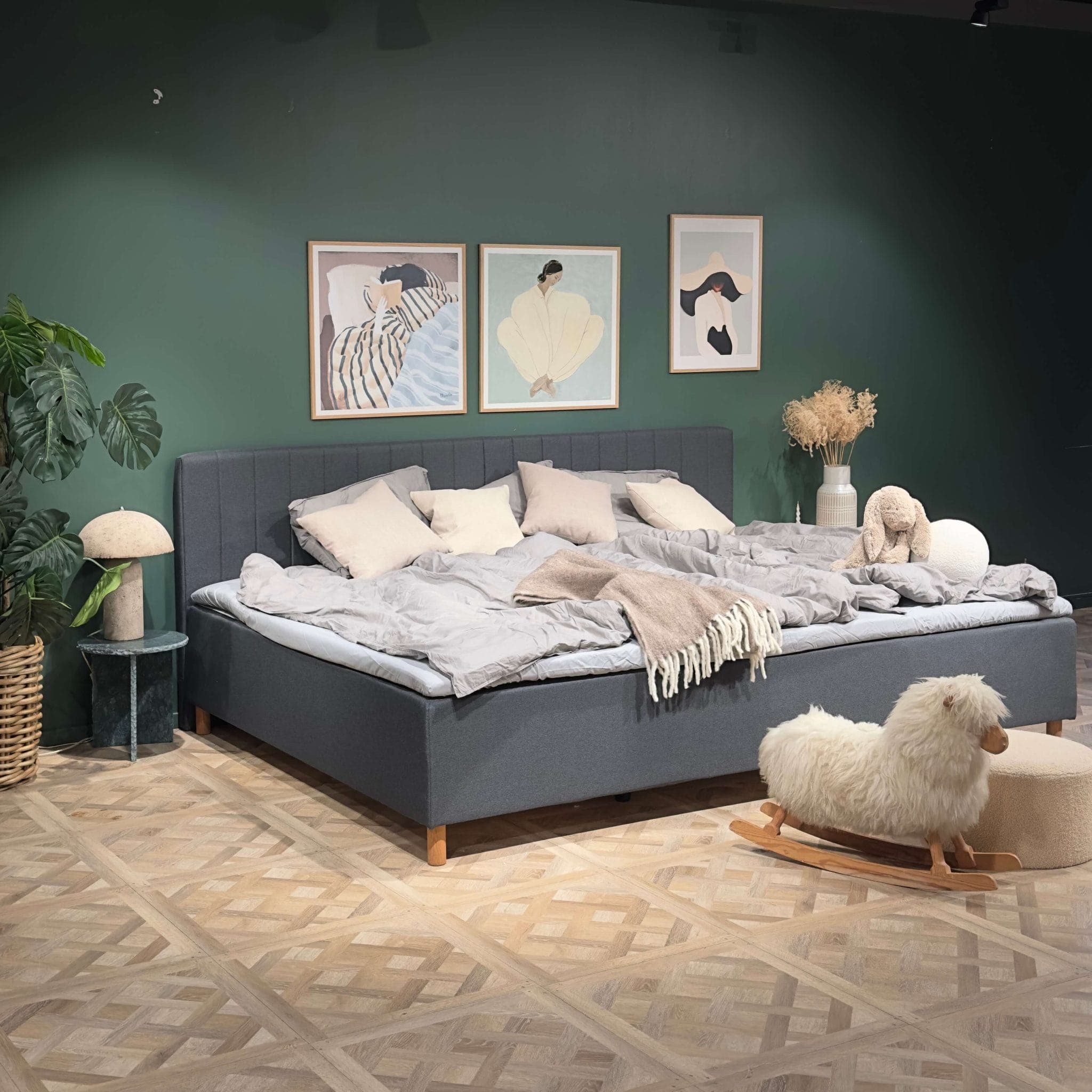Široká ustlaná šedá rodinná postel v ložnici se zelenou stěnou a houpací ovečkou na podlaze