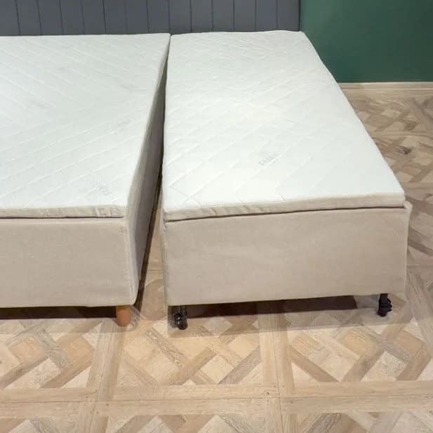 Dva kompletně sestavené moduly postele stojí vedle sebe na světlé dřevěné podlaze a odděluje je úzká mezera. Každý modul má vlastní béžový potah, prošívanou matraci a nohy.