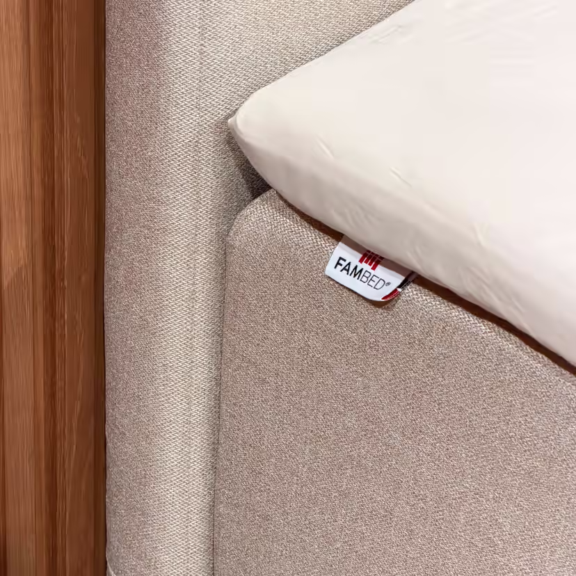 Detail béžového potahu obepínajícího roh postele, se štítkem FAMBED a bílým prostěradlem navrchu